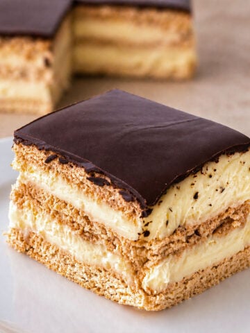 No-bake chocolate eclair dessert slice with creamy custard layers and biscuit base on a plate, topped with smooth chocolate glaze