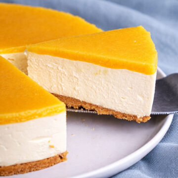 Slice of no-bake orange cheesecake with a glossy orange topping and biscuit crust lifted onto a plate.