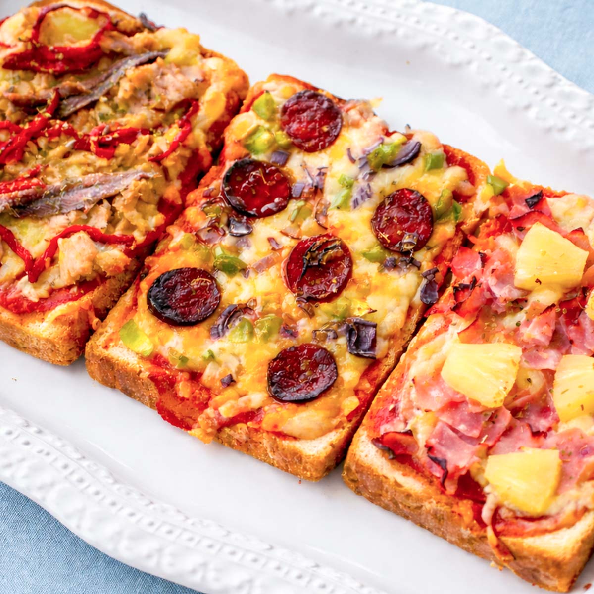 pizza toast with three toppings on sandwich bread, tuna and peppers, chorizo with vegetables, and ham with pineapple, baked with melted cheese on a white plate