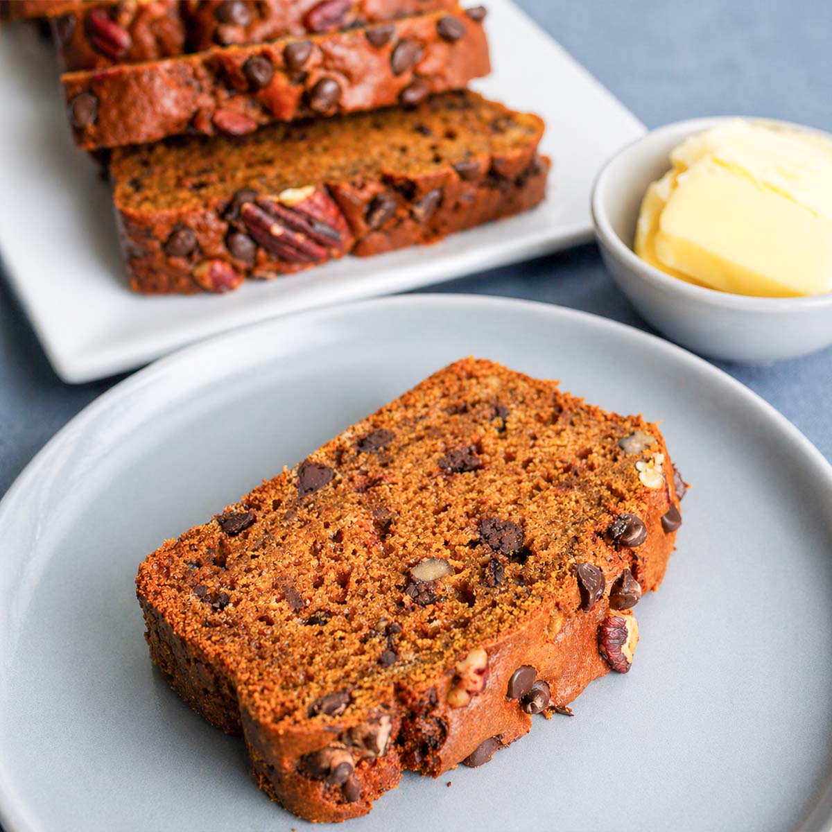 Slice of Pumpkin Bread with Chocolate Chips and Pecans on a plate, with extra slices and a small bowl of butter in the background