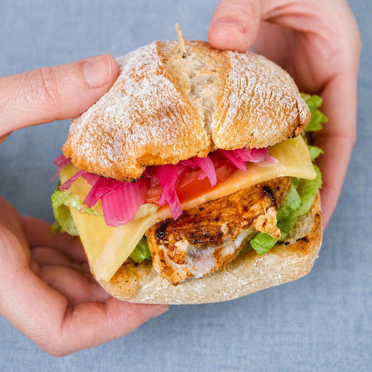 grilled chicken sandwich with pickled red onions cheese lettuce and tomato on ciabatta bread held in hands