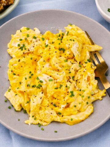 Soft scrambled eggs topped with fresh chives and black pepper on a gray plate with a fork.