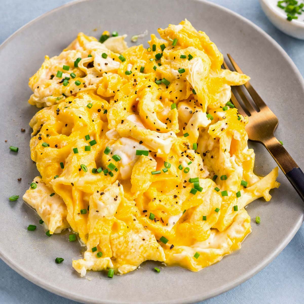Soft scrambled eggs with chives and black pepper served on a gray plate with a fork.