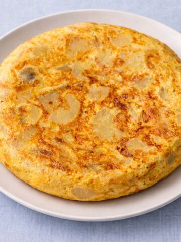 Spanish omelette (tortilla española) with potatoes and eggs served on a white plate against a soft linen blue background.