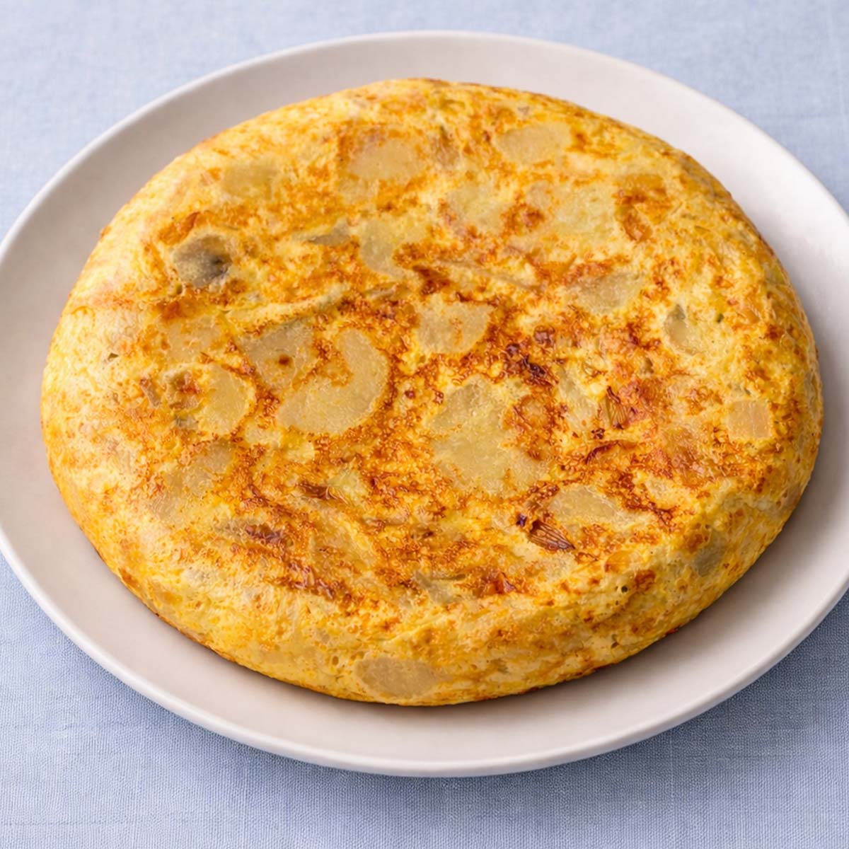 Tortilla Española (Spanish Omelette) with potatoes and eggs served on a white plate against a soft linen blue background.
