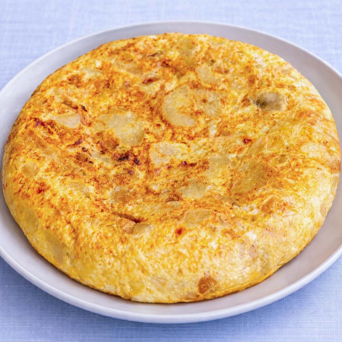 Tortilla Española (Spanish Omelette) made with potatoes and eggs, served whole on a white plate with a soft linen blue background.