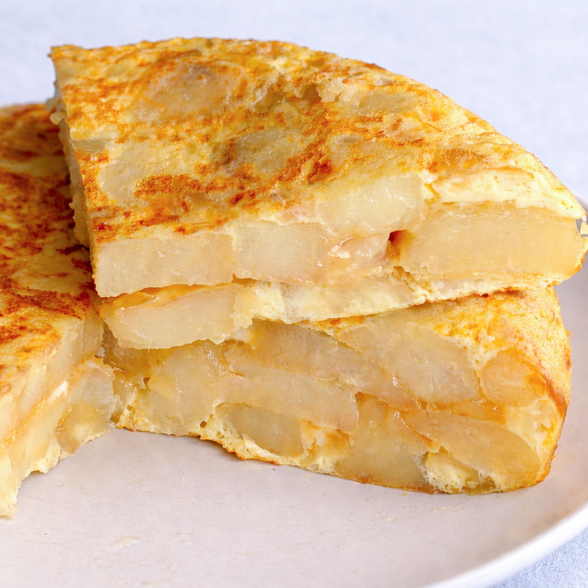 Stacked slices of Spanish omelette (tortilla española) showing layers of tender potatoes and eggs on a white plate against a soft linen blue background.