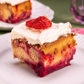 Strawberry shortcut cake slice with creamy frosting and fresh strawberry topping on a white plate, showing layers of vanilla cake and strawberry filling.