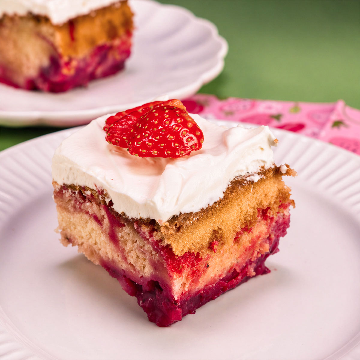 Slice of strawberry shortcut cake with creamy frosting and fresh strawberry on top, served on a white plate with visible layers of vanilla cake and strawberry filling.