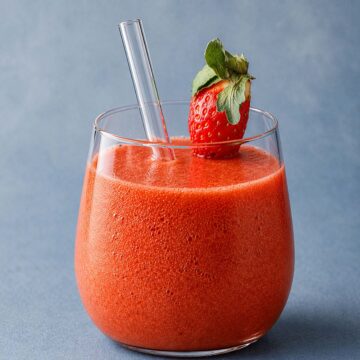 Fresh strawberry mango smoothie in a glass with a strawberry garnish and straw on a muted blue background.