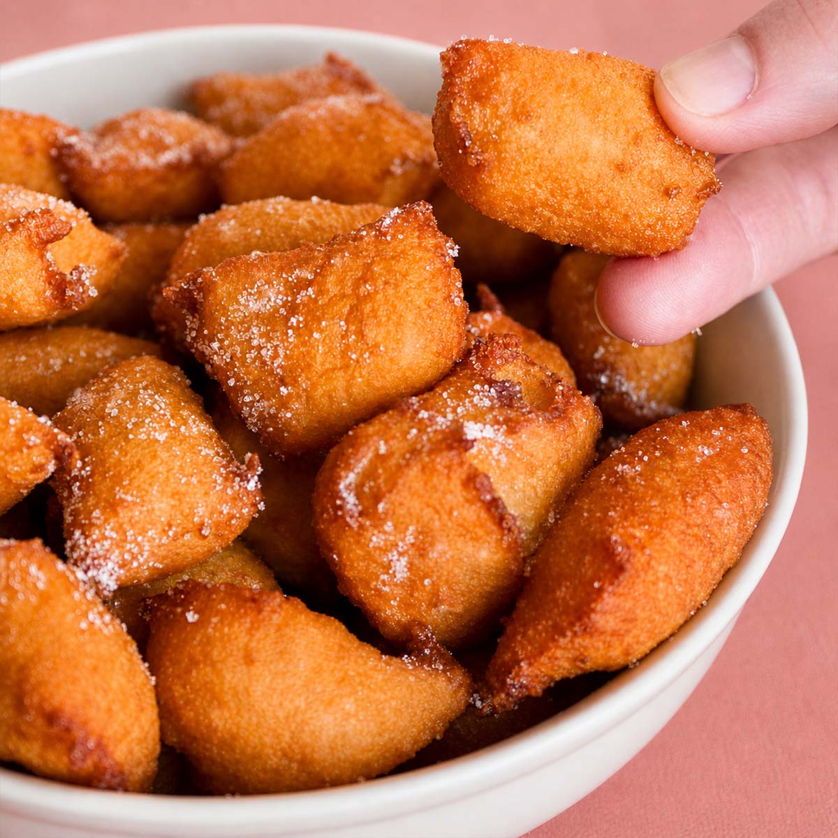 Apple fritters coated in sugar in a bowl, golden and crispy, with a hand picking one up