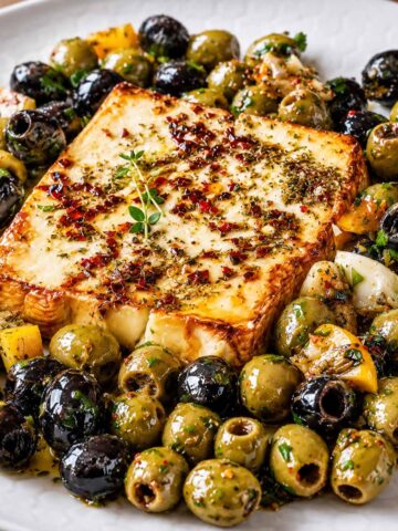 Baked Feta Recipe with Marinated Olives served on a white plate with herbs, chili flakes, and citrus for a Mediterranean appetizer