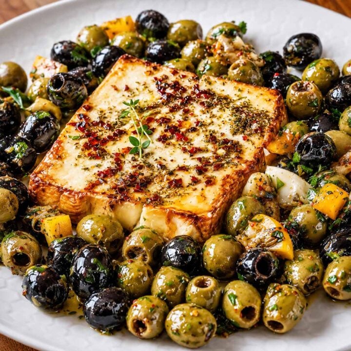 Baked Feta Recipe with Marinated Olives served on a white plate with herbs, chili flakes, and citrus for a Mediterranean appetizer