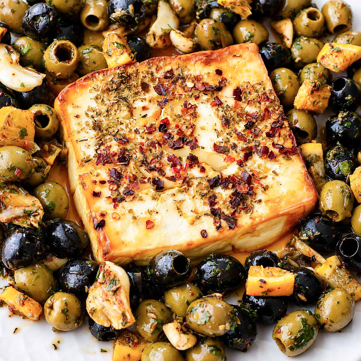 Close-up of Baked Feta Recipe with Marinated Olives topped with chili flakes and herbs, surrounded by citrus-marinated green and black olives.