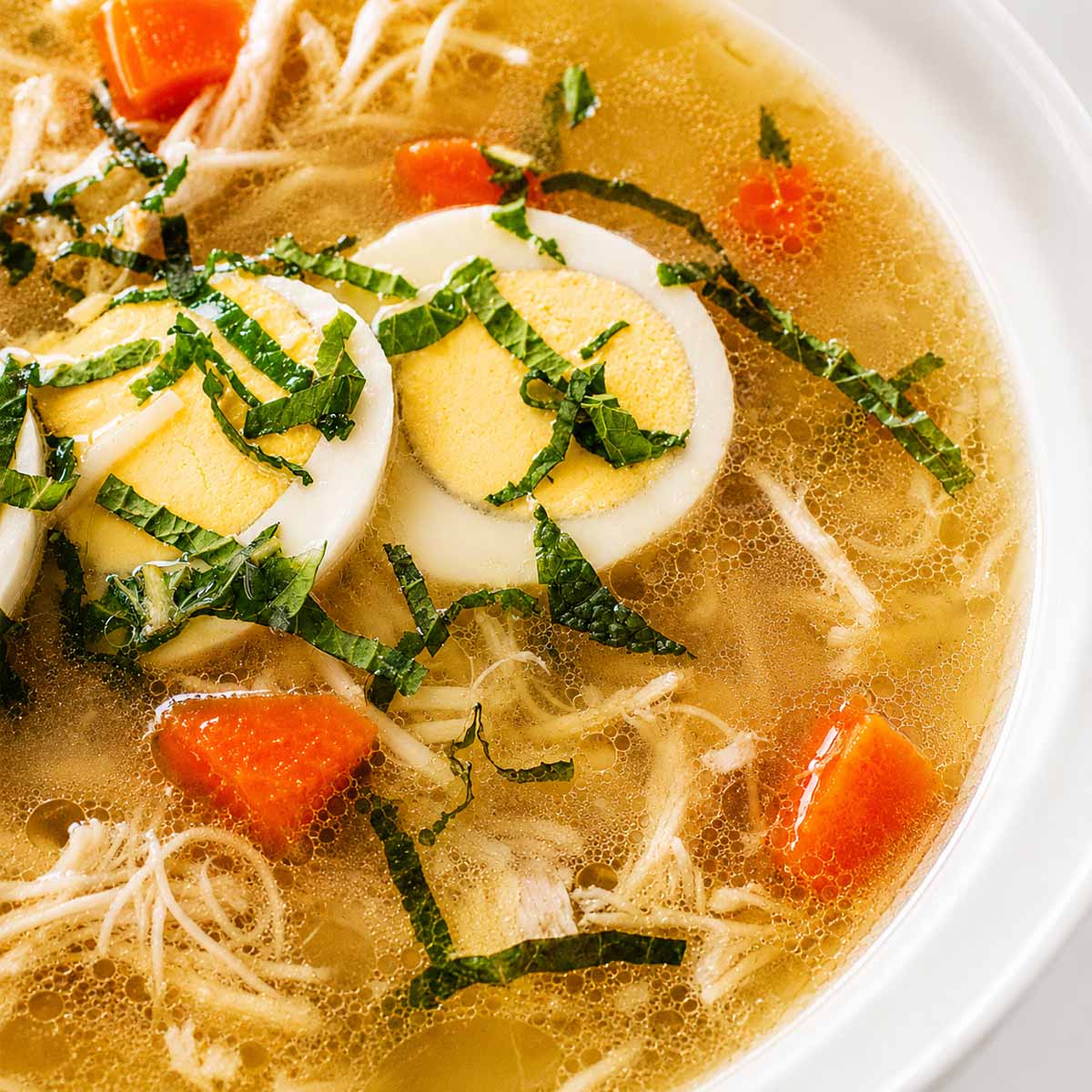 Close up Chicken noodle soup with shredded chicken, carrots, thin noodles, and sliced boiled eggs in a clear golden broth