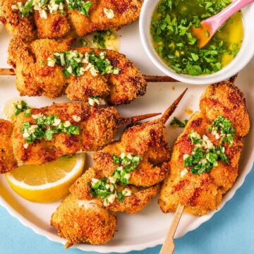 Close-up of crispy lemon garlic butter chicken spiedini skewers with Parmesan breadcrumb coating, topped with chopped garlic and parsley, served with lemon and a bowl of garlic butter sauce.