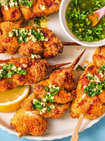 Close-up of crispy lemon garlic butter chicken spiedini skewers with Parmesan breadcrumb coating, topped with chopped garlic and parsley, served with lemon and a bowl of garlic butter sauce.