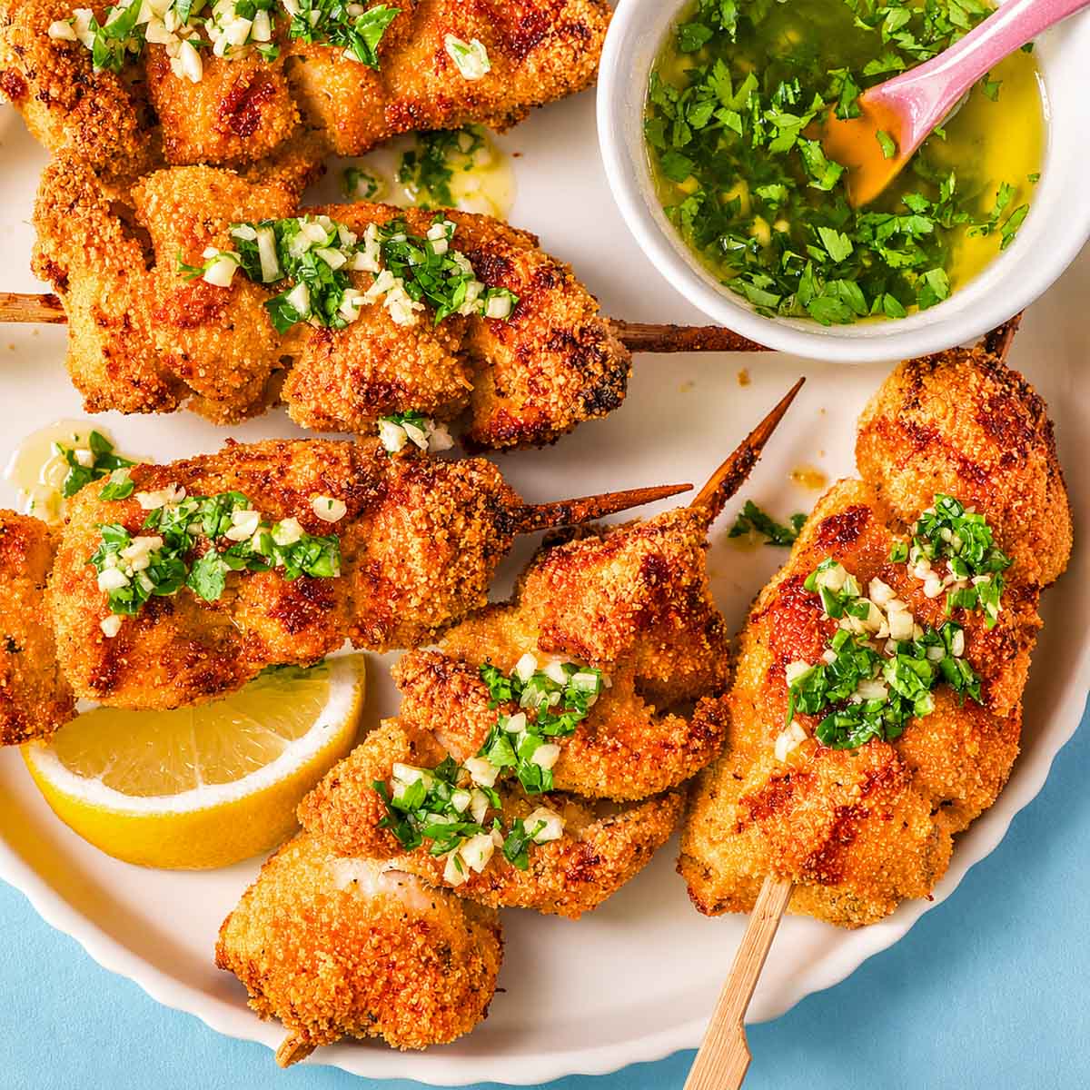 Close-up of crispy lemon garlic butter chicken spiedini with Parmesan breadcrumb coating, topped with chopped garlic and parsley, served with lemon and a bowl of garlic butter sauce.