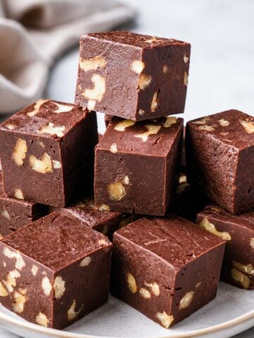 Chocolate walnut fudge cubes stacked on a white plate with a creamy smooth texture and crunchy walnuts inside