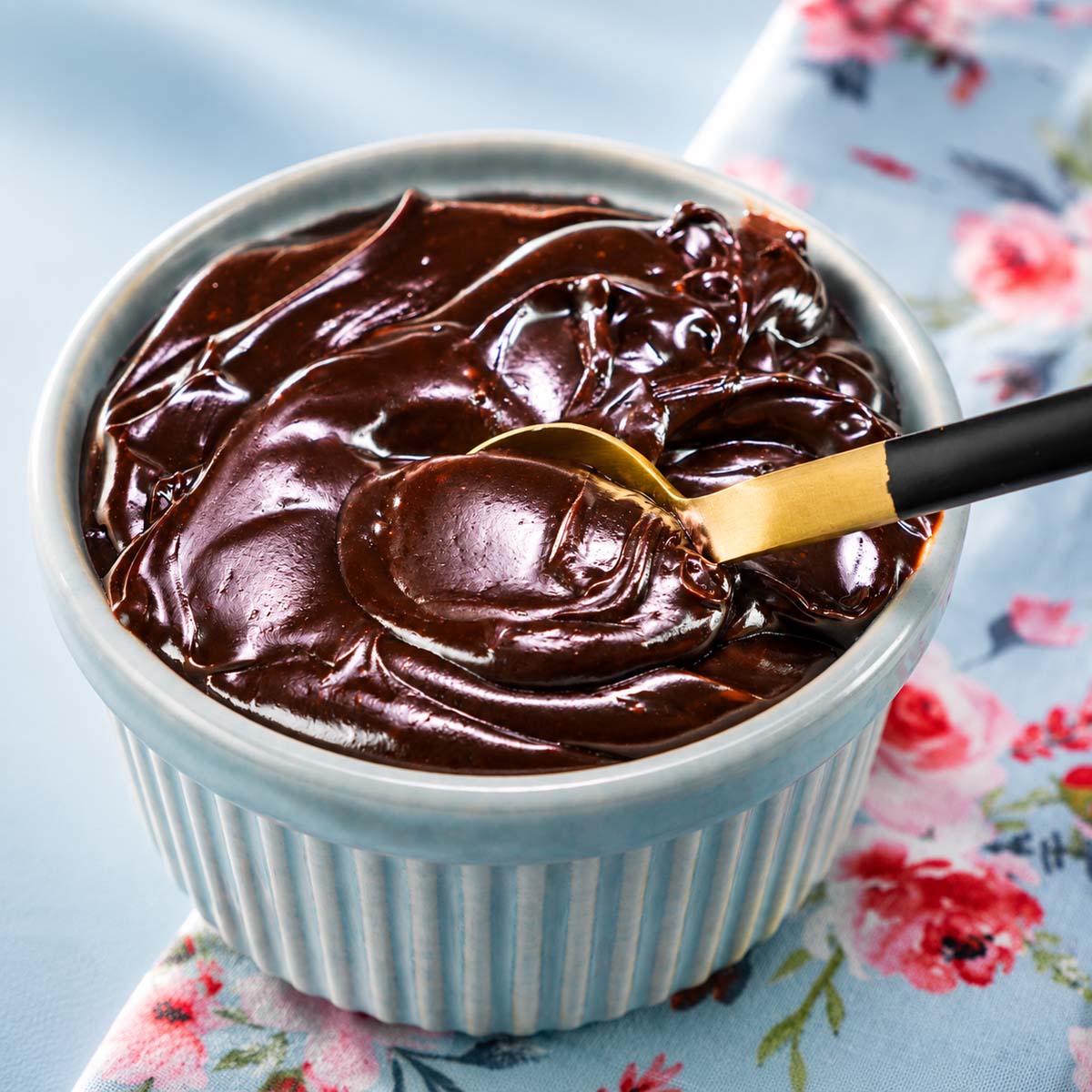 Chocolate frosting with cocoa powder in a blue ramekin with a spoon lifting the glossy creamy frosting