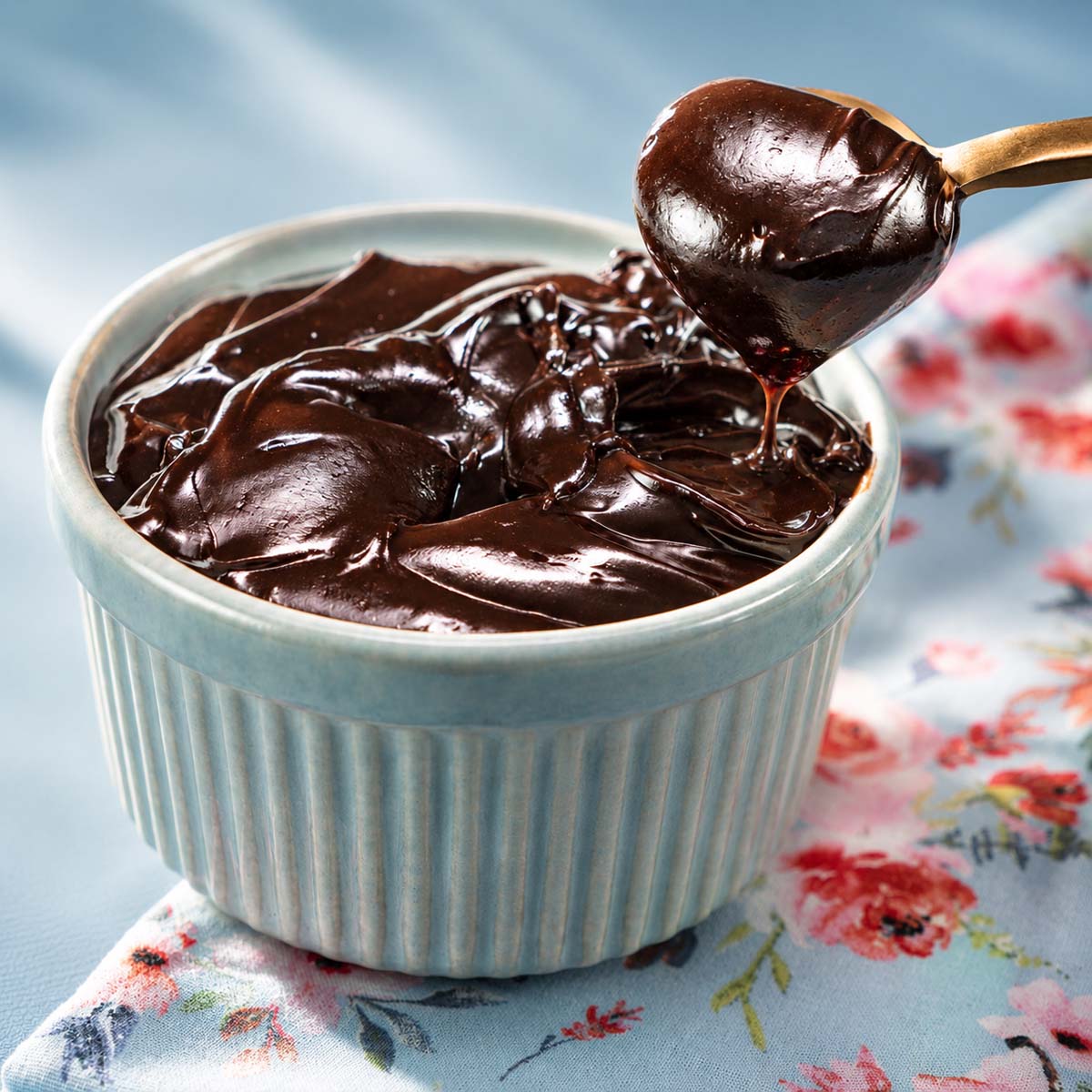 Chocolate frosting with cocoa powder in a blue ramekin with a spoon lifting the glossy creamy frosting