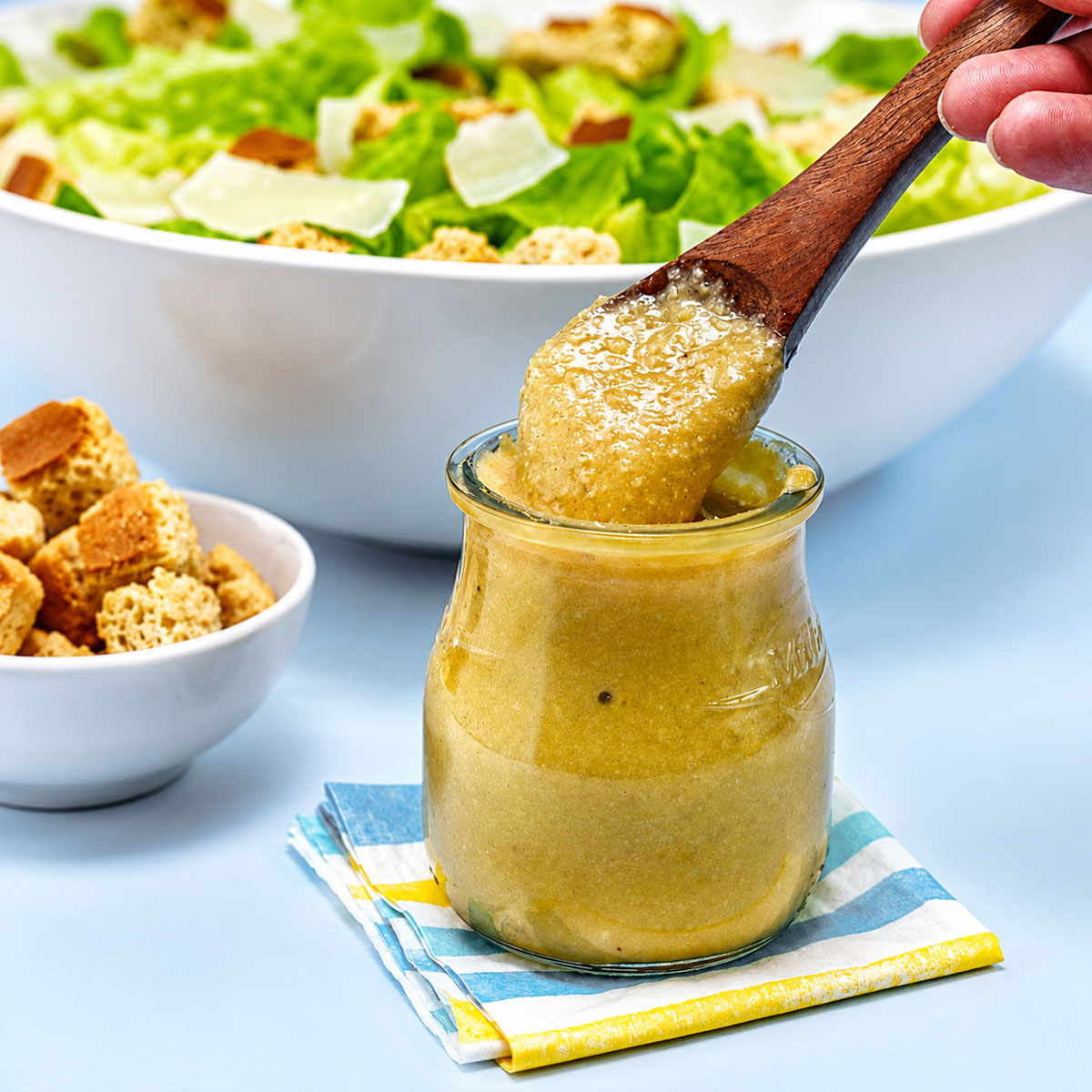 Creamy Easy Caesar Salad Dressing scooped from a glass jar with a wooden spoon, served alongside fresh romaine lettuce, croutons, and Parmesan.