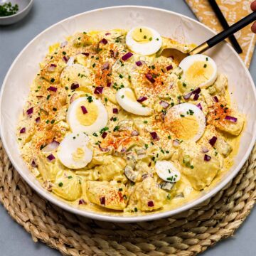 Creamy egg potato salad in a white serving bowl topped with sliced boiled eggs, paprika, red onion, and fresh chives.