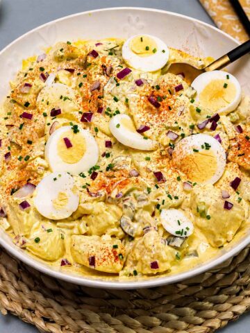 Creamy egg potato salad in a white serving bowl topped with sliced boiled eggs, paprika, red onion, and fresh chives.