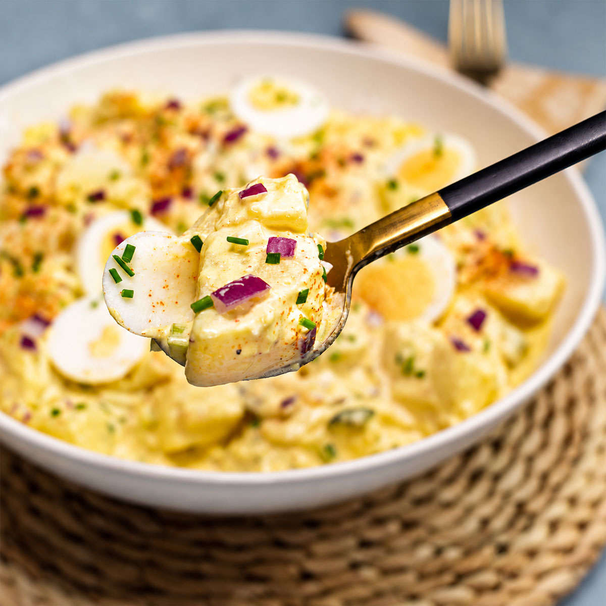 Close-up spoonful of creamy egg potato salad with tender potatoes, boiled egg, red onion, paprika, and fresh chives in a serving bowl.