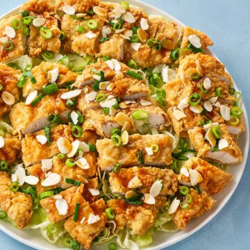 Crispy fried chicken breasts topped with spring onions, almond flakes, and savory sauce on a fresh lettuce platter with a golden crunchy coating.