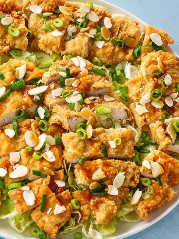Crispy fried chicken breasts topped with spring onions, almond flakes, and savory sauce on a fresh lettuce platter with a golden crunchy coating.