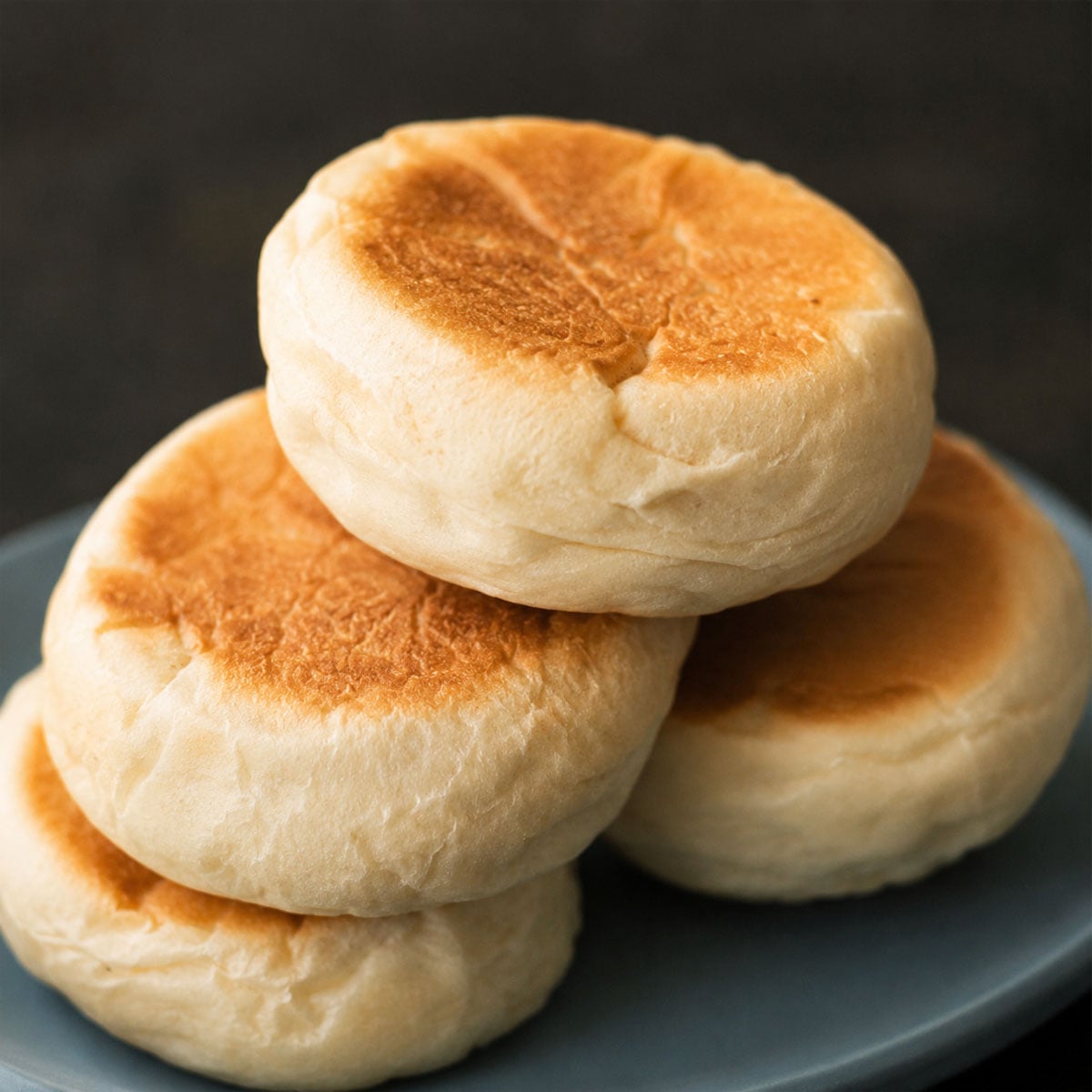 Close up homemade English muffins stacked on a blue plate with golden tops and a soft, fluffy texture, ready for breakfast or brunch.