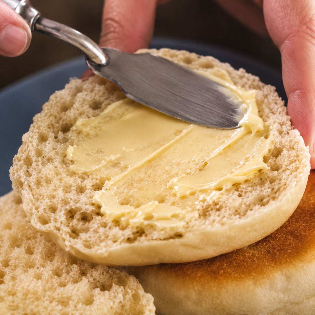Butter being spread on a freshly split homemade English muffin, showing its soft interior, airy texture, and classic nooks and crannies.