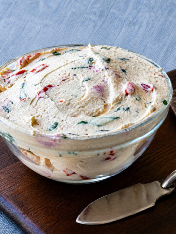 homemade labneh in a glass bowl with herbs and red pepper flakes, served on a wooden board with crackers and a spreader knife