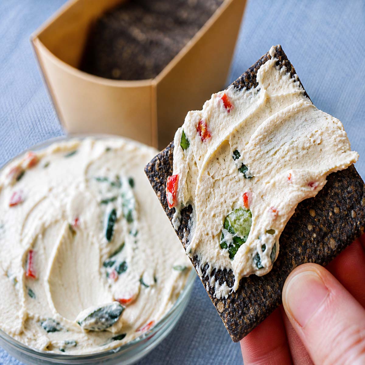 homemade labneh spread on a seeded cracker held by hand, with a bowl of herbed labneh in the background
