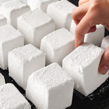 Homemade marshmallows cut into soft cubes and dusted with powdered sugar on a tray, with a hand picking one up