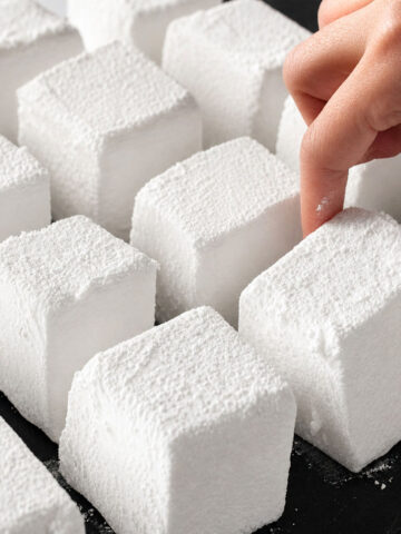 Homemade marshmallows cut into soft cubes and dusted with powdered sugar on a tray, with a hand picking one up
