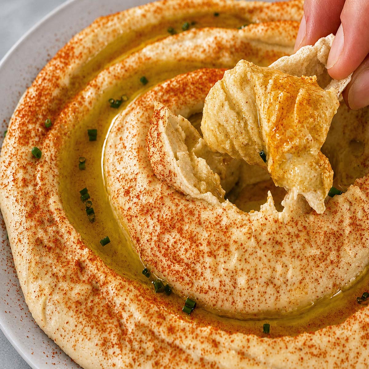 Creamy hummus without tahini swirled with olive oil, paprika, and chives on a plate, with a hand scooping the humus with pita chips
