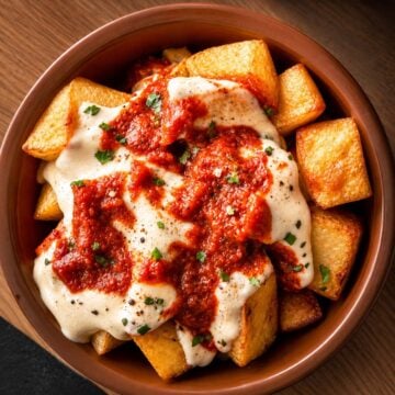 Golden crispy patatas bravas topped with creamy aioli and smoky Spanish brava sauce in a rustic ceramic bowl.