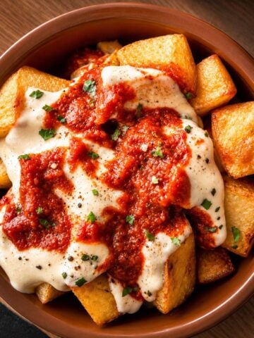 Golden crispy patatas bravas topped with creamy aioli and smoky Spanish brava sauce in a rustic ceramic bowl.