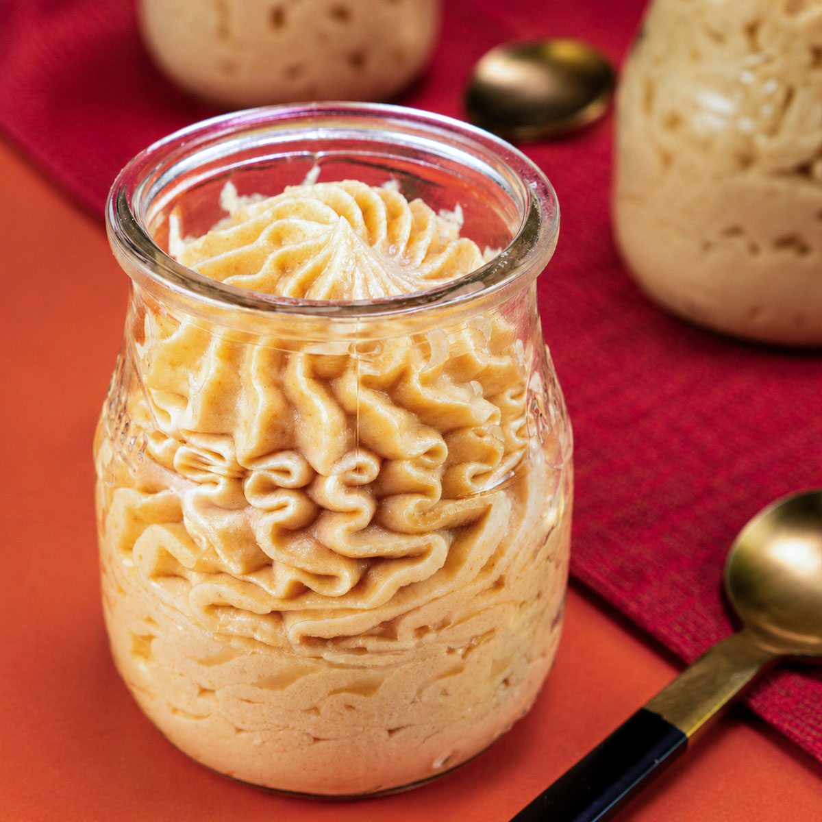 Peanut Butter Mousse in a glass jar with decorative piped swirls, served on a red background with gold spoons and additional mousse jars.