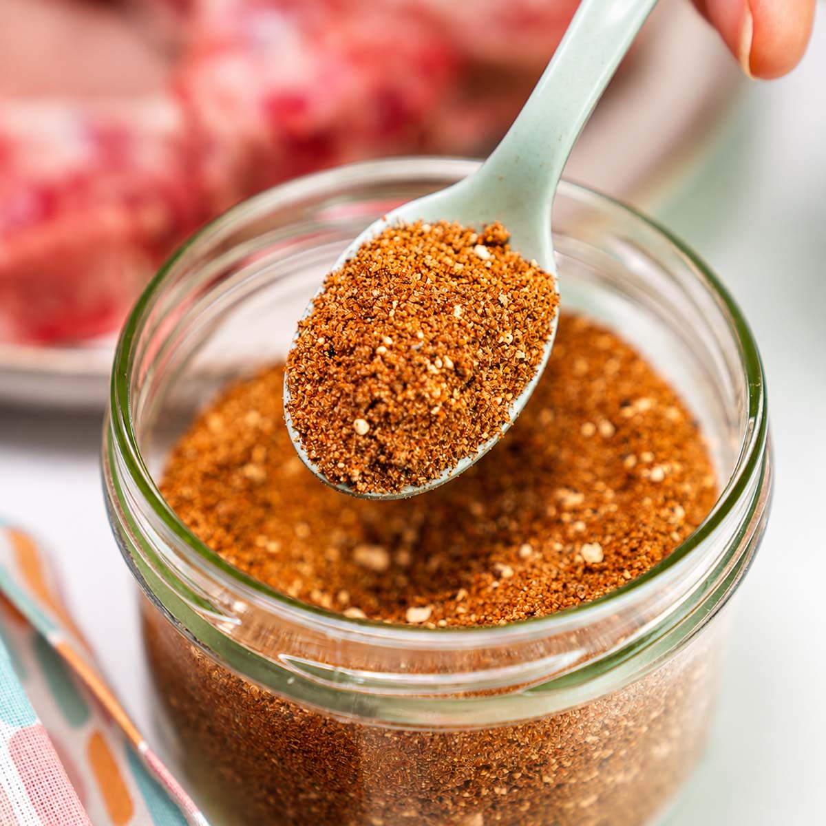 close up Pork chop seasoning in a glass jar with spoon beside raw pork chops on a plate