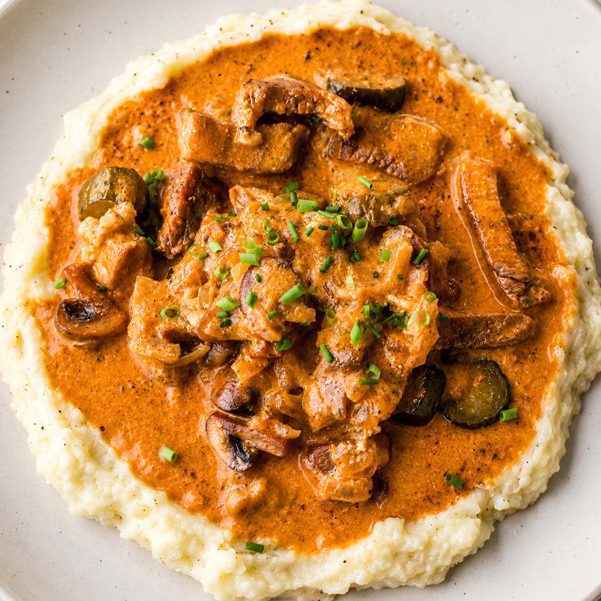 Creamy pork stroganoff served over mashed potatoes with mushrooms and pickles in a rich sauce