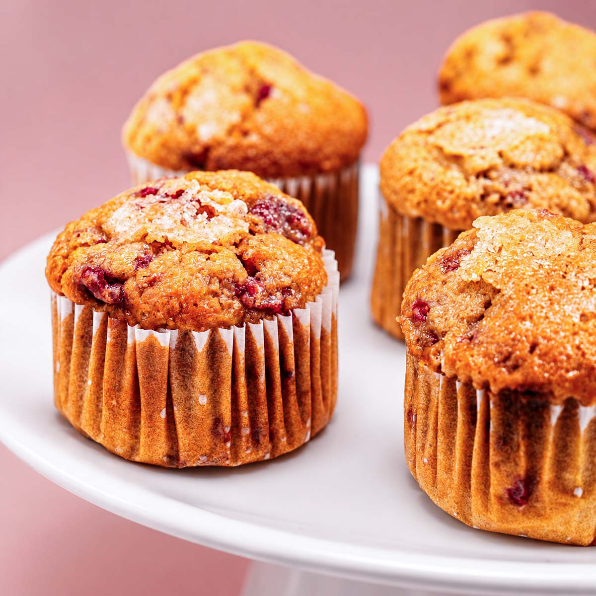Close-up raspberry muffins with golden sugary tops and berry-filled centers served on a white cake stand
