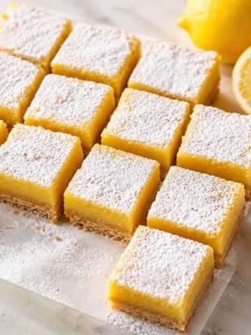 Soft lemon squares dusted with powdered sugar on a marble surface with fresh lemon slices and a buttery crust visible in each creamy layer