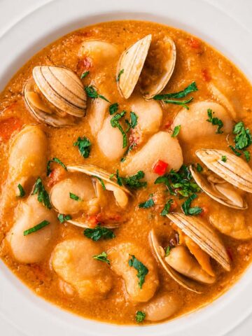 Spanish Bean Soup with Clam (Alubias Blancas con Almejas) served in a white bowl, featuring creamy white beans, tender clams in shell, and a rich broth topped with fresh herbs
