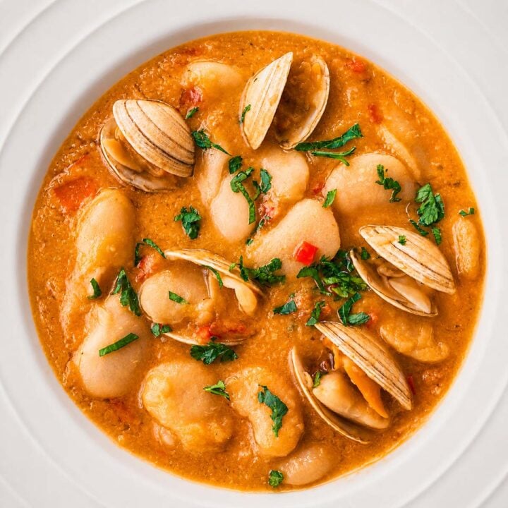 Spanish Bean Soup with Clam (Alubias Blancas con Almejas) served in a white bowl, featuring creamy white beans, tender clams in shell, and a rich broth topped with fresh herbs