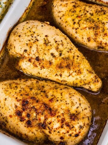 Close-up of baked Italian dressing chicken breast with golden seasoning and herby pan juices in a baking dish.