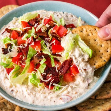 Cracker dipping into creamy BLT dip topped with crispy bacon, diced tomatoes, shredded romaine, and cheddar cheese.