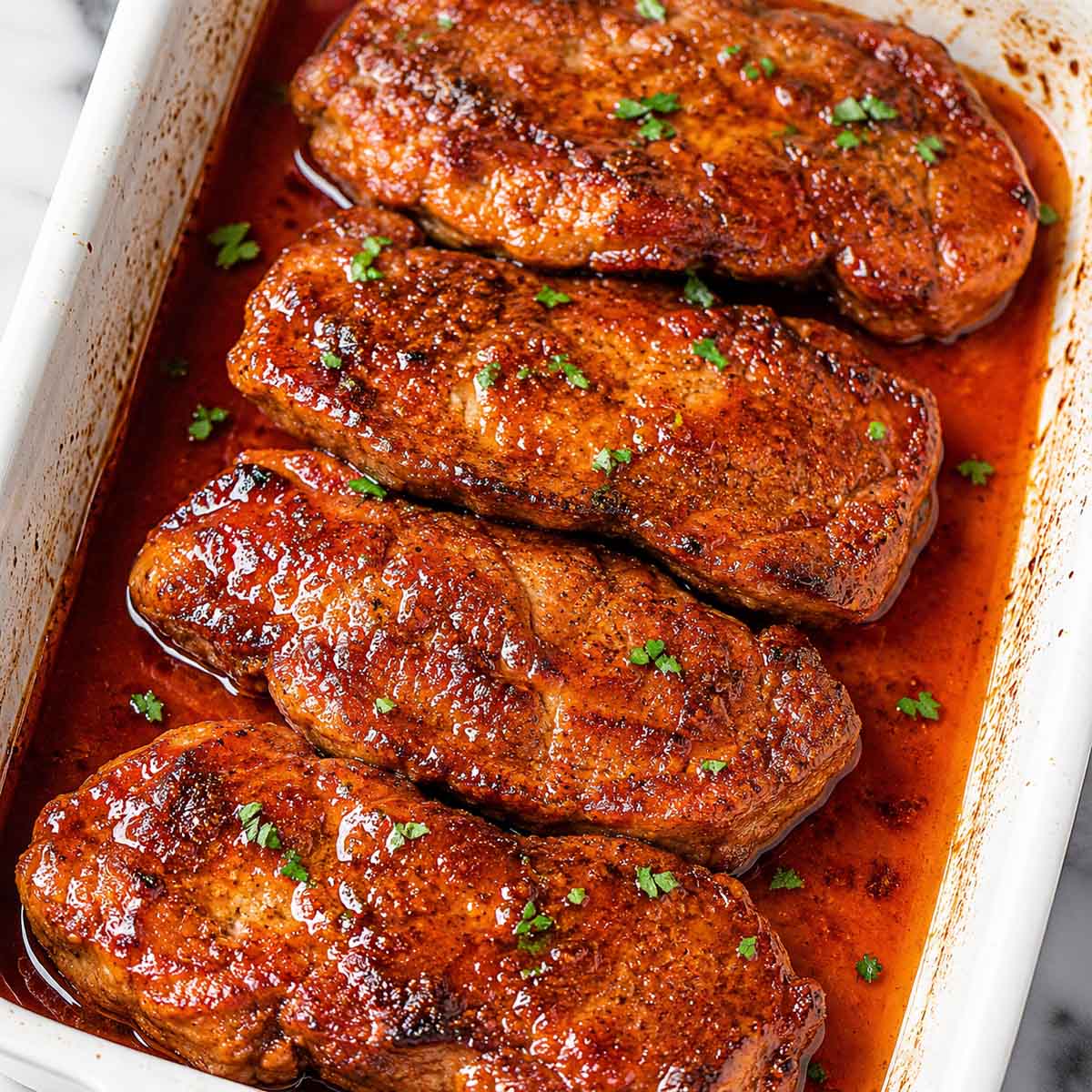 Baked pork steaks in a rich reddish sauce, garnished with chopped parsley and cooked until tender in the oven.